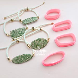 May include: Four adjustable bracelets featuring green and white marbled clay plaques on white cord with gold-tone metal accents, accompanied by four pink plastic cutters in oval and rectangular shapes. The text Sylhouette Studios is visible.