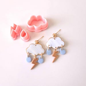 May include: A pair of handmade earrings featuring a white cloud with a marbled effect, gold lightning bolts, and blue raindrops. The earrings are made with polymer clay and are lightweight and comfortable to wear.