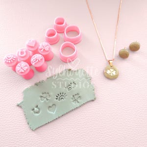 May include: A collection of pink clay tools, including circle cutters and stampers with heart, horseshoe, and clover designs. A necklace with a clover pendant and acorn earrings are also displayed on a pink surface.