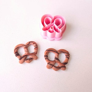 Puede incluir: Dos galletas de pretzel de chocolate con glaseado marrón y naranja. También se muestra un cortador de galletas en forma de corazón de plástico rosa.