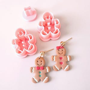 May include: Two gingerbread man earrings with red bows and gold hooks. The earrings are made of polymer clay and are a light brown color. The earrings are next to two pink gingerbread man cookie cutters.