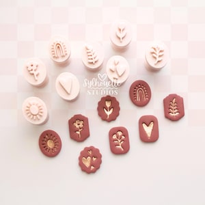 Op de afbeelding: Een verzameling lichtroze ronde kleistempels met reliëfmotieven van bloemen, harten, regenbogen en bladeren, naast verschillende donkere terracotta-kleurige kleistukken met goudkleurige afdrukken van dezelfde botanische en geometrische vormen.