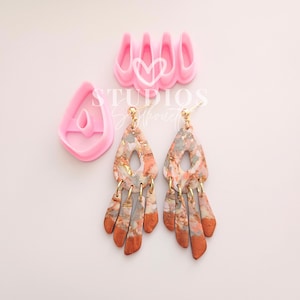 Puede incluir: Cortadores de pendientes de arcilla polimérica rosa y un par de pendientes hechos a mano. Los pendientes tienen forma de lágrima con una parte superior dorada y un diseño multicolor gris, naranja y dorado. El texto "STUDIOS Silhouette" es visible.