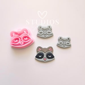 Può includere: Un tagliabiscotti rosa a forma di faccia di procione accanto a tre biscotti grigi a forma di faccia di procione. Le facce dei procioni hanno gli occhi chiusi e nasi rosa. Il tagliabiscotti ha la forma di una faccia di procione.