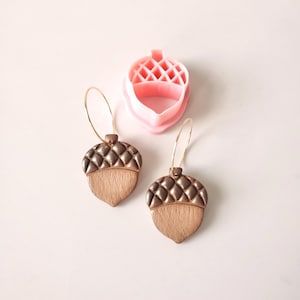 May include: A pair of gold hoop earrings with acorn-shaped charms. The charms are made of brown clay with a textured surface and a gold finish. The earrings are shown next to a pink plastic acorn-shaped cookie cutter.