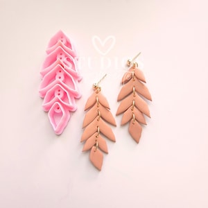 May include: Pink polymer clay earring cutter next to a pair of peach-colored leaf earrings with gold-colored hardware. The earrings are made of multiple leaf-shaped pieces connected by small gold rings. The cutter is a stack of leaf shapes.