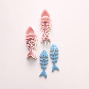 Può includere: Due paia di orecchini a forma di lisca di pesce blu con anelli dorati. Gli orecchini sono realizzati in pasta polimerica e hanno la forma di lisca di pesce. Gli orecchini sono mostrati accanto a due tagliapasta a forma di lisca di pesce in plastica rosa.