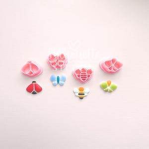 May include: A selection of colourful cookie cutters shaped like a ladybug, butterfly, bee, and flowers. The cutters are in shades of red, blue, yellow, green, and pink. The background is a soft pink.