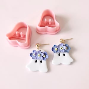 May include: A pair of white ghost-shaped earrings with purple flower accents. The earrings are made of polymer clay and have gold ear wires. The earrings are shown with two pink plastic molds for making the earrings.