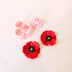 May include: Two red polymer clay poppy flowers with black centers and yellow accents.  A set of three pink plastic flower cutters.