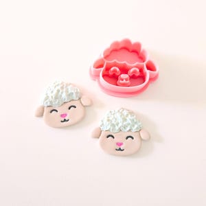 May include: Two pink and white fondant sheep faces with smiling faces and pink noses. A pink plastic cookie cutter in the shape of a sheep face.