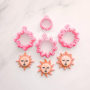 May include: A selection of pink sun-shaped cookie cutters and decorated biscuits. The cutters are in various styles, including a ring and sunburst shapes. The biscuits are orange with a face design, featuring closed eyes and a small mouth.