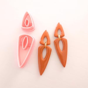 May include: A pair of terracotta-colored earrings with a geometric design. The earrings are made of a thick, glossy material and have a gold-colored accent in the center. The earrings are shown next to two pink plastic molds.