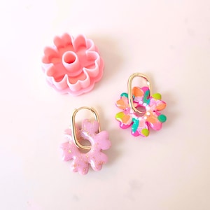 May include: Two pairs of gold hoop earrings with pink and multi-colored flower charms. The charms are made of polymer clay and have a painted design. The earrings are displayed on a white surface.