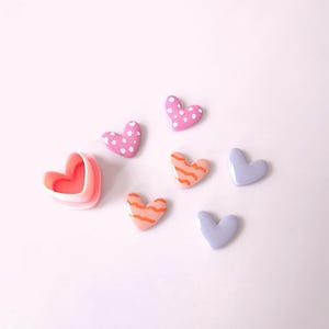 以下が含まれることがあります： 様々な色と模様のハート型のボタン7個セット。ボタンはピンク、パープル、オレンジで、白い水玉模様、ストライプ、無地です。