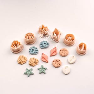 Puede incluir: Una colección de cortadores de galletas de plástico en color melocotón, azul y blanco y formas de arcilla a juego con varios diseños de conchas marinas y estrellas de mar. El set incluye varias formas moldeadas y cortadores con el texto Sylhouette Studios.