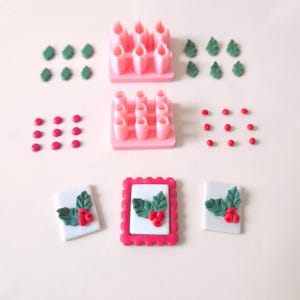 Puede incluir: Un conjunto de moldes de silicona rosa para hacer decoraciones navideñas. Los moldes incluyen hojas de acebo, bayas y velas. También hay decoraciones prefabricadas en la imagen, incluyendo hojas de acebo, bayas y una rama de acebo enmarcada.