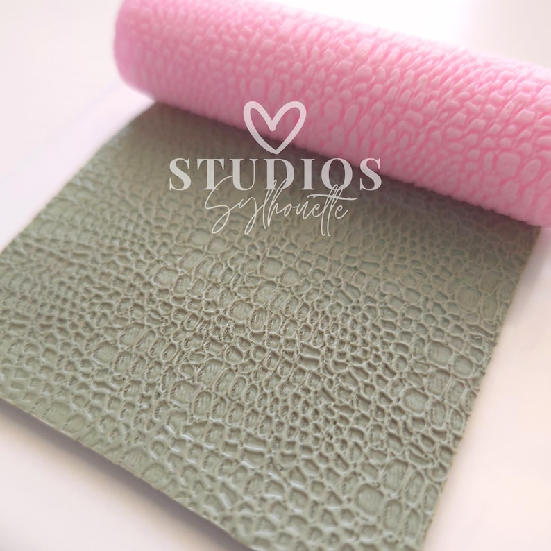 May include: A rolled pink textured material rests above a flat, textured, sage green material. The materials have a pattern that resembles reptile skin. The text "STUDIOS Syhouette" is printed on the sage green material.