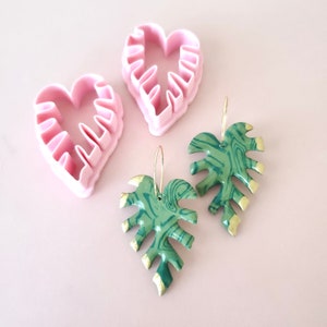 Puede incluir: Dos pendientes en forma de hoja verde y dorada con un patrón de mármol. Los pendientes cuelgan de aros dorados. También hay dos cortadores de galletas en forma de corazón rosa en la imagen.