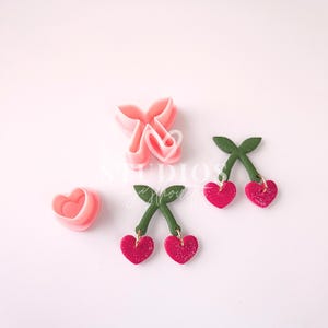 Puede incluir: Un juego de tres cortadores de galletas de plástico rosa en forma de cerezas. Los cortadores se utilizan para crear galletas de cereza en forma de corazón. Los cortadores están sobre una superficie blanca.