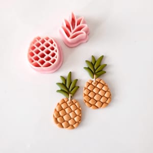 Könnte beinhalten: Zwei Paare von Ananas-Ohrringen mit einem strukturierten Ananas-Design und grünen Blättern. Die Ohrringe sind aus Polymer Clay gefertigt und haben eine hellbraune Farbe.
