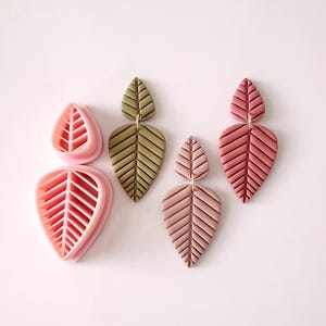 May include: Three pairs of leaf-shaped earrings. One pair is green, one pair is pink, and one pair is a pink clay mold. The earrings are made of polymer clay and have a textured surface.