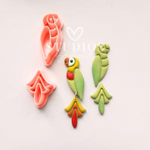 May include: A set of polymer clay cutters for creating parrot shapes. The cutters are pink and include a parrot, a tail, and a leaf. There is also a finished parrot made of green, yellow, and red polymer clay.