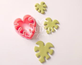 Puede incluir: Cortador de galletas de corazón rosa con diseño de hoja de monstera. Se muestran tres formas de hojas de monstera verdes junto al cortador.