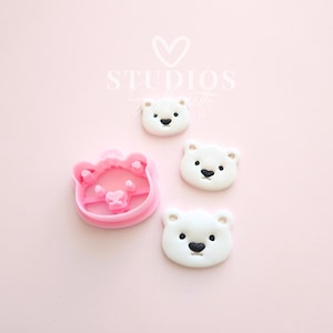 May include: A pink bear-shaped cookie cutter and three white bear-shaped biscuits with black noses and eyes, on a pink background. The text "STUDIOS" and "Sashalette" is in the upper left corner.