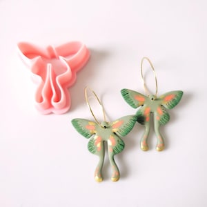 Puede incluir: Pendientes de polilla hechos a mano en verde y dorado con aros dorados. Los pendientes tienen un diseño único con detalles en naranja. También se ve un cortador de arcilla rosa con forma de mariposa.