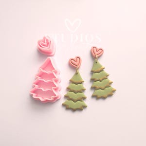 May include: Pink and green Christmas tree-shaped earrings and cookie cutters. The earrings have a heart-shaped top and gold accents. The cookie cutters are pink and shaped like a heart and a Christmas tree. The background is a light pink.