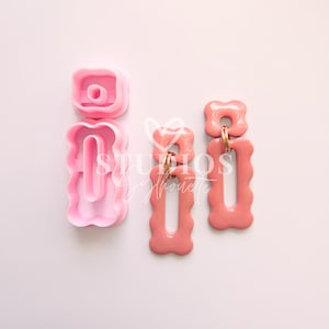 May include: Pink polymer clay earring cutters and finished earrings. The cutters are in the shape of a square with a circle in the middle and a rectangle with a wavy edge. The earrings are a coral color with gold-colored earring hooks.