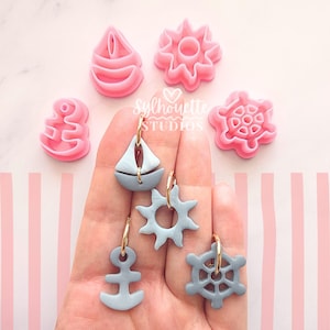 May include: A hand displays nautical-themed earrings and pink clay cutters. The earrings feature a sailboat, sun, anchor, and ship's wheel in light blue with gold hoops. Pink cutters shaped like a boat, sun, anchor, and ship's wheel are also visible.