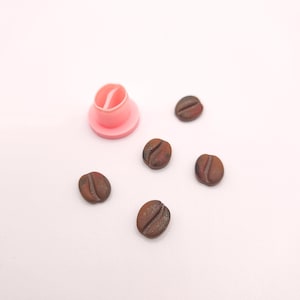 May include: A pink coffee bean-shaped cookie cutter next to five brown coffee bean-shaped cookies. The cookies have a textured surface and are scattered on a white background. The cookie cutter is made of plastic.