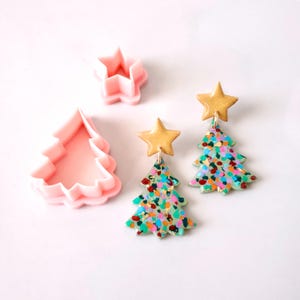 以下が含まれることがあります： 斑点模様のペイントデザインが施された、緑と金のクリスマスツリーのイヤリングのペア。イヤリングはポリマークレイ製で、トップに金の星チャームが付いています。