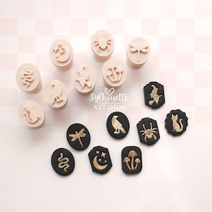 May include: A collection of small oval and shaped stamps featuring embossed designs of a snake, moon, spider, dragonfly, cat, raven, witch, and mushrooms. Some stamps are light beige, while others are black with gold-colored embossed details.