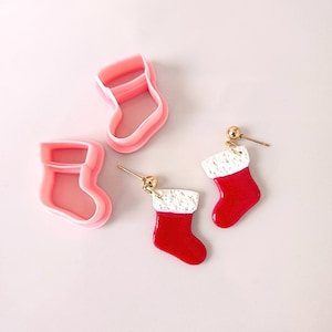 Puede incluir: Dos pares de pendientes de calcetines de Navidad rojos y blancos. Un par está hecho de arcilla polimérica y el otro es un cortador de galletas.