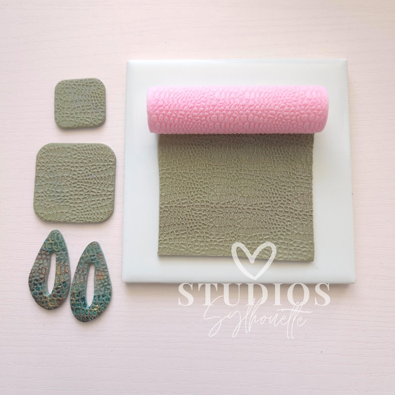 May include: An overhead shot displays crafting supplies on a white surface. A pink textured roller sits atop a textured green square. Two smaller green squares and two teal teardrop-shaped earrings are also present. The text "STUDIOS Silhouette" is visible.