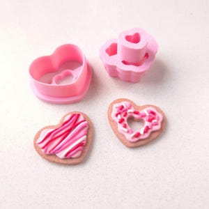 May include: Two pink heart-shaped cookies, one with pink and white stripes and the other with pink frosting and sprinkles, and two pink heart-shaped cookie cutters.