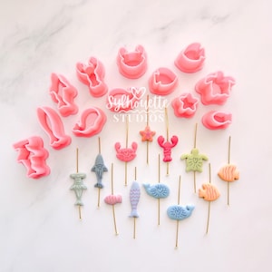 Può includere: Un assortimento di formine per biscotti e decorazioni a tema marino, di colore rosa. Le formine hanno varie forme e le decorazioni raffigurano creature marine come pesci, granchi e aragoste. Il testo "Silhouette Studios" è visibile.