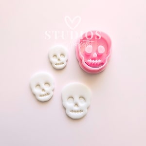 May include: A pink skull-shaped cookie cutter next to three white skull-shaped cookies. The cookie cutter is made of pink plastic. The cookies are white and have detailed facial features. The background is a light pink color.