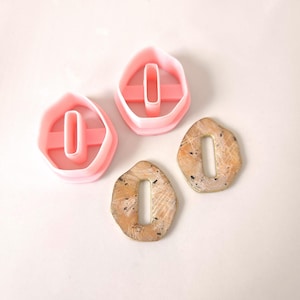 May include: Two pink plastic cookie cutters shaped like a rounded rectangle with a rectangular hole in the centre. Two gold-coloured metal cookie cutters shaped like a rounded rectangle with a rectangular hole in the centre.