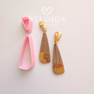 May include: Earrings with a wooden teardrop shape, accented with gold leaf and a textured gold section at the bottom. Two pink teardrop-shaped cutters are also in the image. The earrings are hanging from gold-colored hooks.