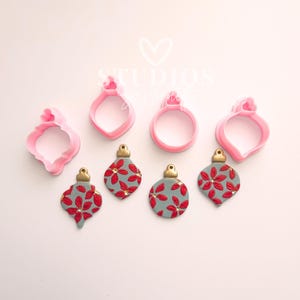 May include: Pink plastic ornament-shaped cookie cutters and decorated ornaments. The ornaments are gray with red floral designs and gold tops. The cookie cutters are in various shapes, including teardrop and round.