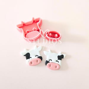 May include: Two pink plastic cookie cutters shaped like cow heads, and two fondant cow heads with black and white spots.