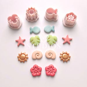 May include: A set of 12 colorful fondant cutters and toppers in a beach theme. The cutters are pink and white plastic and include shapes like a starfish, seashell, sun, fish, and flower. The toppers are made of fondant and are in various colors, including pink, blue, green, gold, and peach.