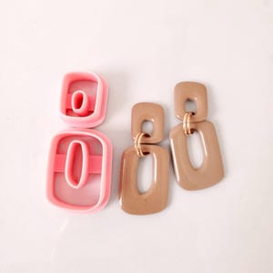 May include: A pair of brown resin earrings with gold accents. The earrings are shaped like a rectangle with a smaller rectangle cut out of the center. The earrings are shown next to two pink plastic cookie cutters.