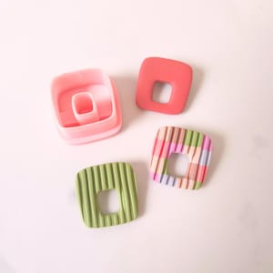 Puede incluir: Tres pendientes cuadrados de arcilla polimérica con un agujero en el centro. Los pendientes son de color rosa, verde y con estampado de cuadros multicolores. También se muestra un molde de plástico rosa.