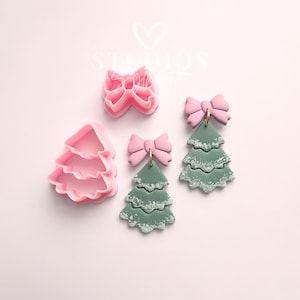 以下が含まれることがあります： ピンク色のリボンとクリスマスツリー型のクッキーカッターとイヤリング。クッキーカッターはピンク色で、イヤリングはピンクのリボンが付いた緑色です。イヤリングには金色のフープが付いています。背景は薄いピンク色です。