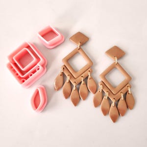 Puede incluir: Un par de pendientes colgantes de color dorado con un diseño de doble cuadrado y abalorios en forma de hoja. Los pendientes están hechos de arcilla polimérica y se muestran junto a cortadores de plástico rosa.